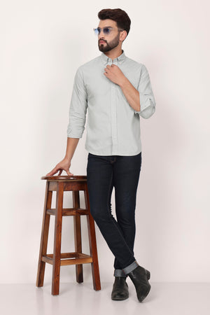 A man seated on a wooden stool wearing sunglasses, a light-colored casual shirt, and black trousers.