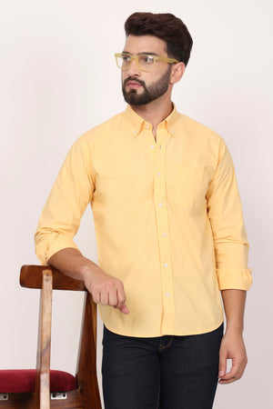 A man wearing a yellow casual shirt with down buttons, paired with black trousers, sitting in front of a chair.